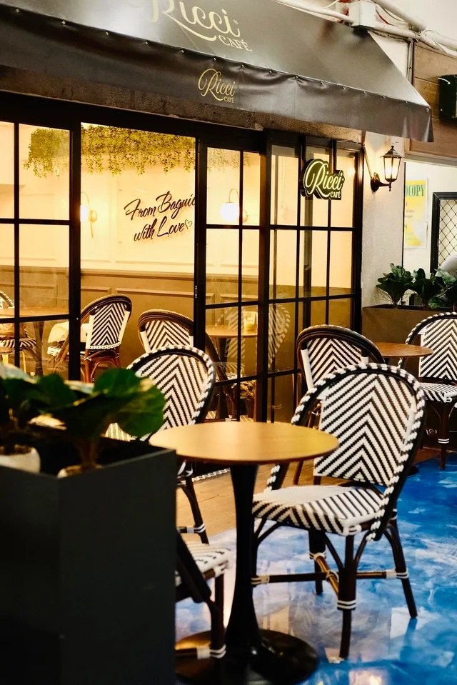 The Ricci Café storefront at Porta Vaga, with its awning and neon Ricci Café sign lit up in the evening, bistro chairs and tables set out front, and a neon script sign reading From Baguio with Love in the window